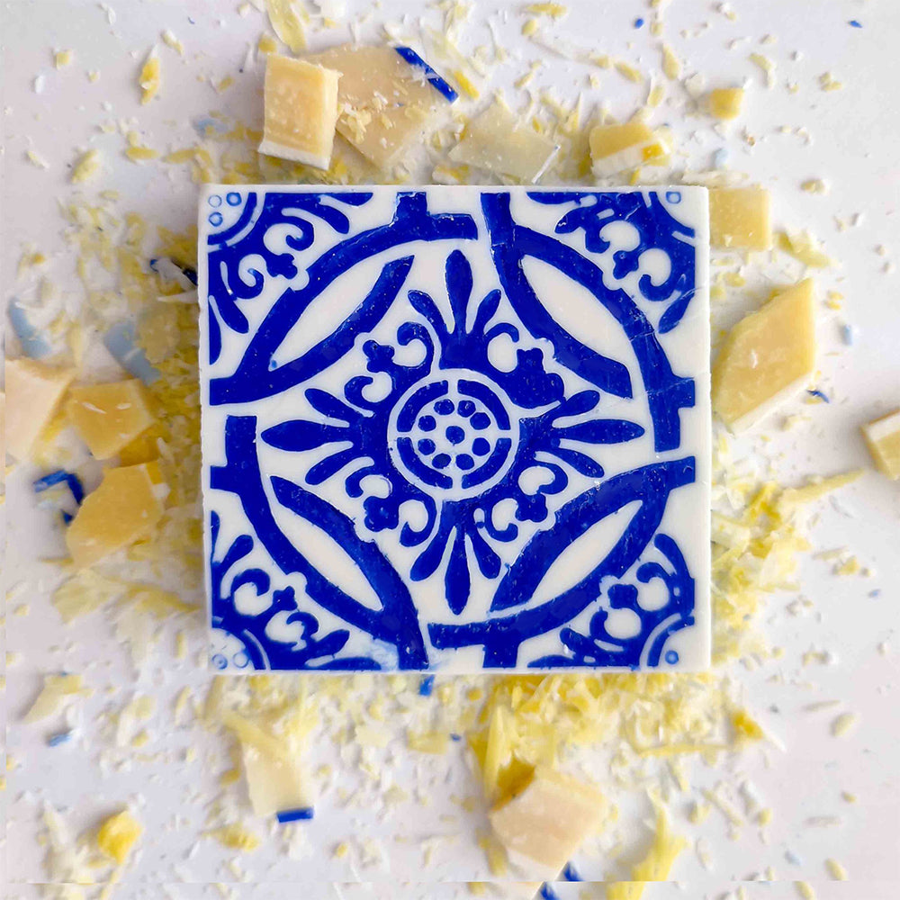 The Lisbão Pastel de Azulejo is a square white handmade Portuguese tile soap with an intricate blue floral design, displayed on a light surface with scattered yellow and white chocolate shavings.