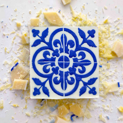 The Lisbão Pastel de Azulejo is a square white Portuguese tile soap with a blue ornate floral and geometric design, displayed among grated cheese and cheese chunks on a white surface.