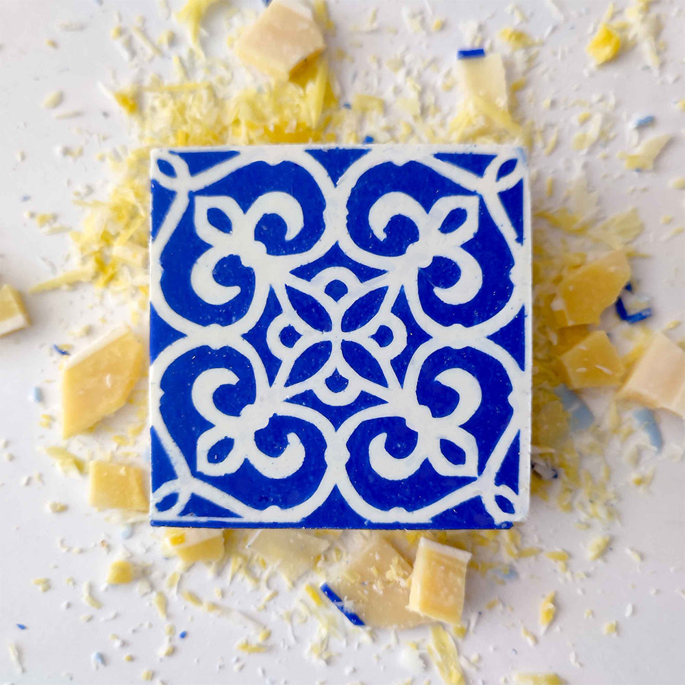 The Lisbão Pastel de Azulejo soap features an intricate blue and white floral tile design, displayed on a white surface with scattered white and yellow cheese shavings and chunks.