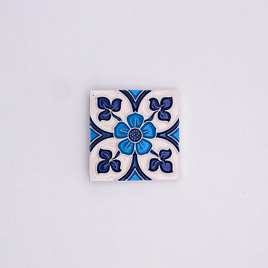 A decorative Tejo Shop Royal Blue Small Ceramic Tile featuring a symmetrical floral design in shades of blue and white, centered with a raised blue button-like detail, against a plain light background.