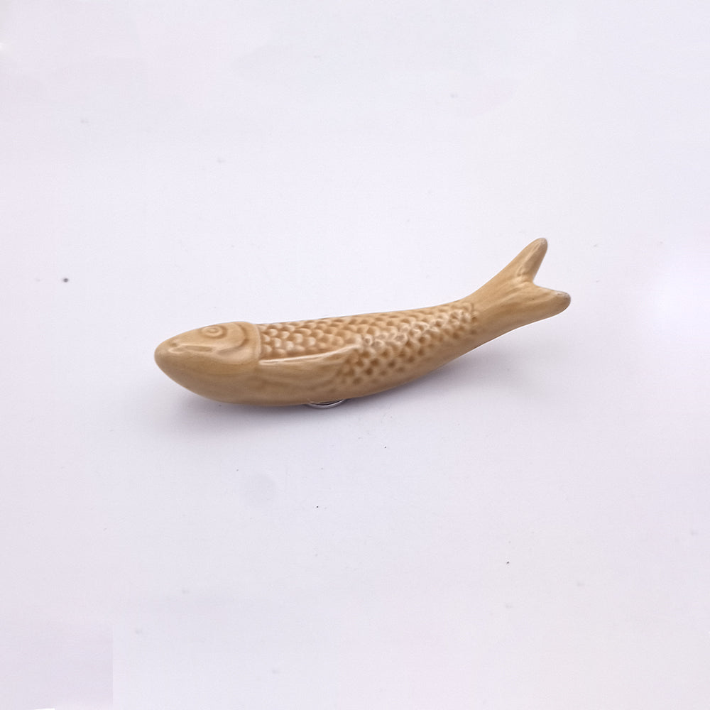 A small ceramic item from Tejo Shop, the Ceramic Sardine Fridge Magnet, resembles a simple fish shape with minimal detailing on its fins and scales. Evoking a classic Portuguese sardine fridge magnet, this neutral beige-colored fish is displayed on a plain white background, making it an elegant piece of kitchen decor.