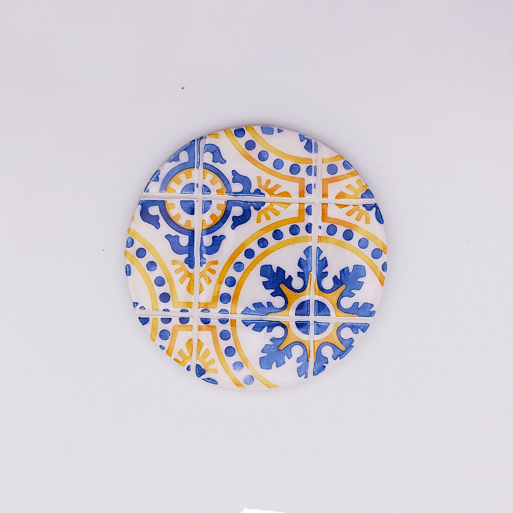 The **Island of Madeira Ceramic Tile Cup Pad** by **Tejo Shop** features a mosaic design with blue and yellow motifs on a white background. The pattern includes intricate floral and geometric shapes, resembling traditional tile artwork, adding artisanal charm to any setting.