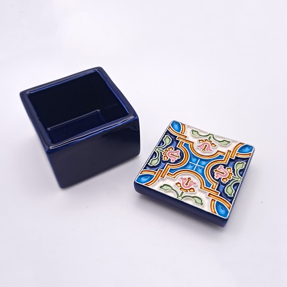 A small, square, dark blue Jerónimos Ceramic Box by Tejo Shop with a fitted lid sits on a white background. The lid features a colorful geometric pattern with shades of blue, pink, green, and cream, forming an intricate design. This unique addition is open, revealing an empty interior that exudes elegance and warmth.