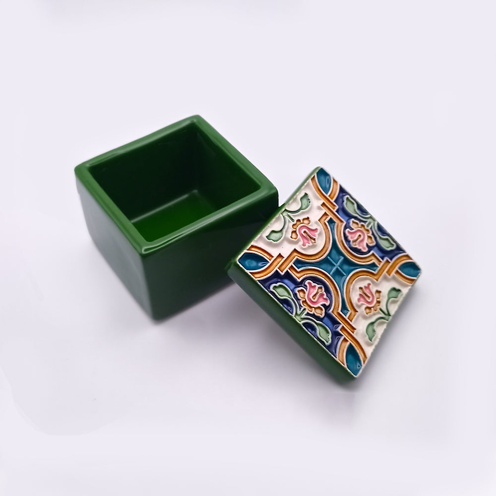 A small, square green box with an open lid, reminiscent of a Lisbon Ceramic Box by Tejo Shop. The lid is decorated with colorful patterns featuring blue, green, orange, and pink tiles in intricate designs. The interior of the box is plain green, and the backdrop is a white surface.