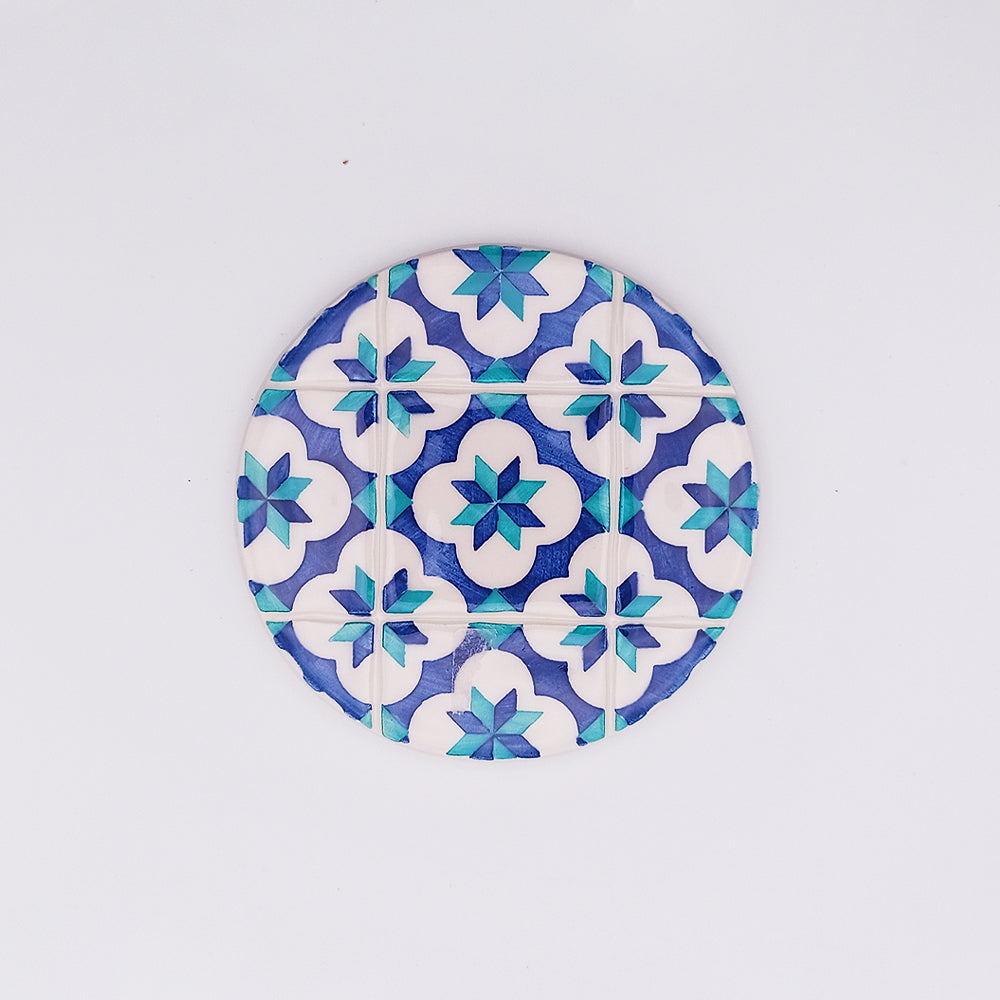 A unique Lisbon Ceramic Tile Cup Pad featuring a blue and white geometric pattern by Tejo Shop. The hand-painted design showcases alternating 8-point stars and white flower-like shapes set on a blue grid. This ceramic cup pad is placed against a plain white background, highlighting its intricate detail.