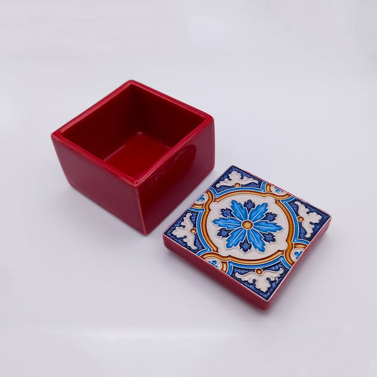 A small, square, red Monsaraz Ceramic Box by Tejo Shop with an open lid placed beside it. The lid features an intricate blue, orange, and white floral pattern. The background is a plain, white surface.