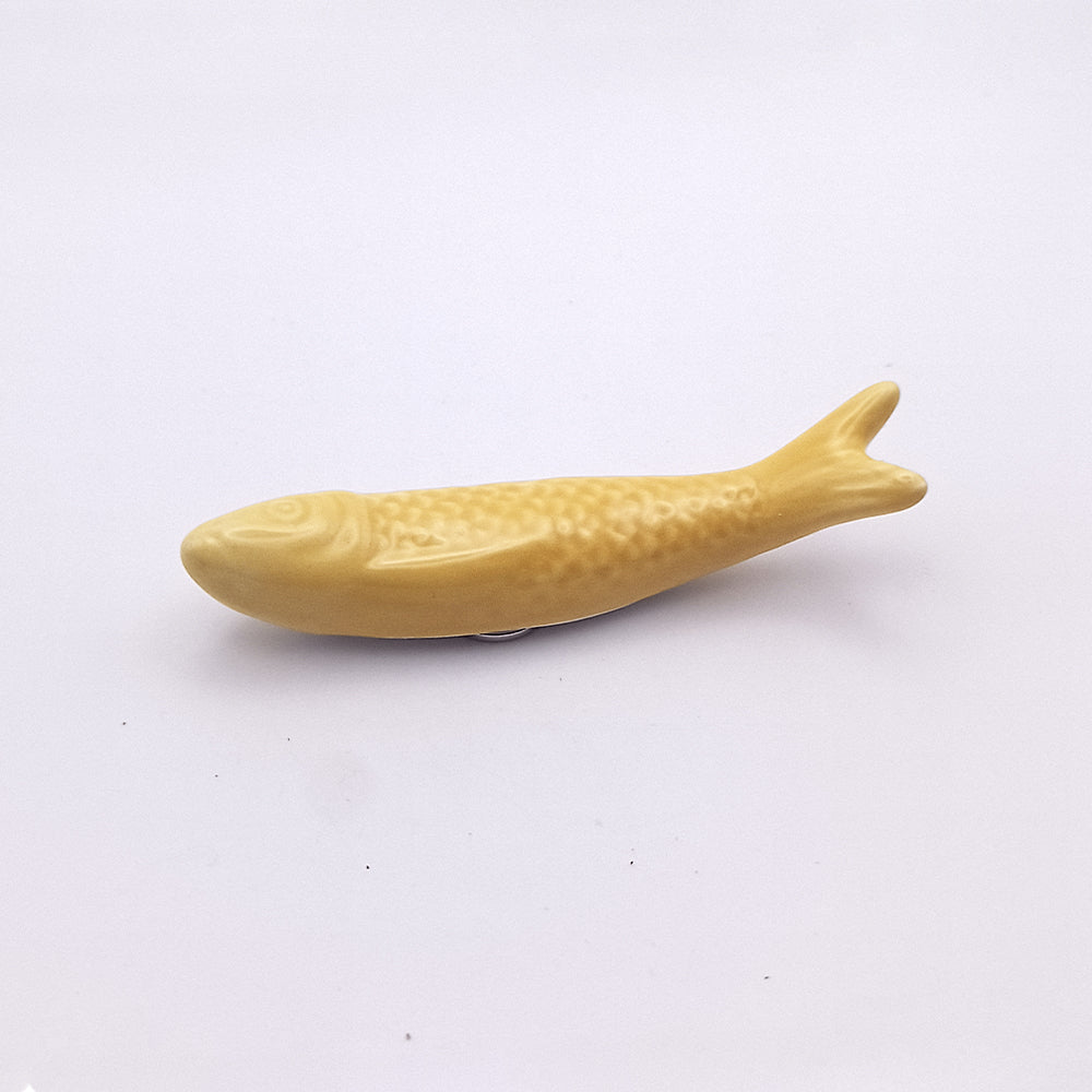 A small, yellow Ceramic Sardine Fridge Magnet from Tejo Shop lies on a plain white background. The sardine has a simple, smooth design with a slight texture to represent scales, embodying a classic piece of Portuguese kitchen decor.