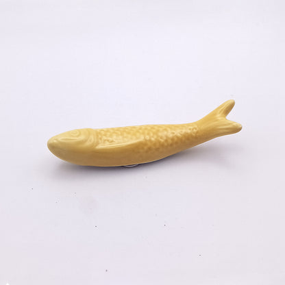 A small, yellow Ceramic Sardine Fridge Magnet from Tejo Shop lies on a plain white background. The sardine has a simple, smooth design with a slight texture to represent scales, embodying a classic piece of Portuguese kitchen decor.