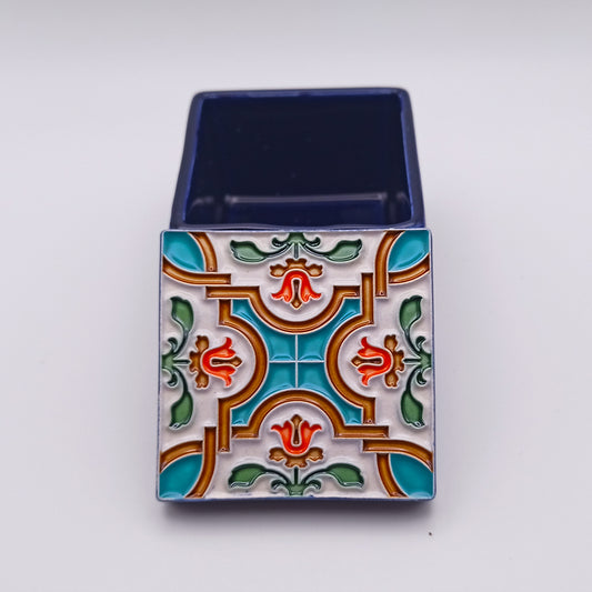 A square, ornately decorated jewelry box with a multicolored, floral and geometric design on the lid. The design features patterns in teal, orange, red, and green. The lid is open, revealing a deep blue interior. This Nazareth Ceramic Box by Tejo Shop is a timeless allure and charming addition to any collection.