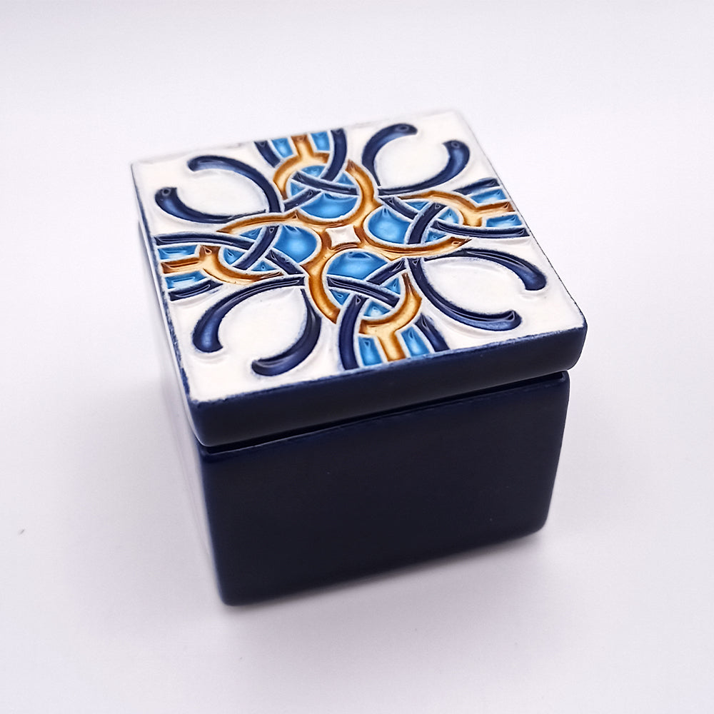 A square, dark blue luxury ceramic box with a detachable lid. The lid features an intricate, colorful geometric pattern in blue, white, and gold. The ornate design consists of interlacing curves and shapes, showcasing expert craftsmanship in its symmetrical and eye-catching decoration. This is the Óbidos Ceramic Box by Tejo Shop.