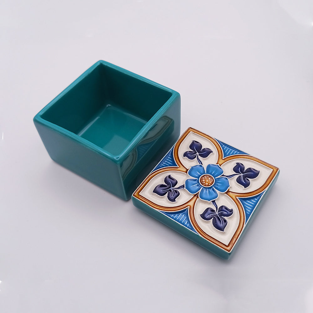 A small, square, teal ceramic box with its lid off. The Pico Islands Ceramic Box by Tejo Shop features an intricate design with a central blue flower surrounded by blue curved shapes and gold accents on a white background. The box's interior is plain teal, emphasizing the hand-painted artistry of the exterior.