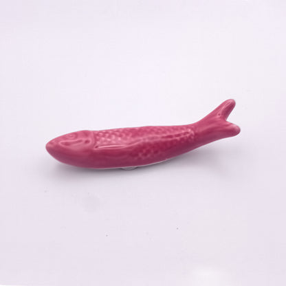 A small, pink Ceramic Sardine Fridge Magnet from Tejo Shop, shaped like a fish and set against a plain white background. This Portuguese icon features simple details, including a textured body and a slightly open mouth, perfect for adding charm to your kitchen decor.