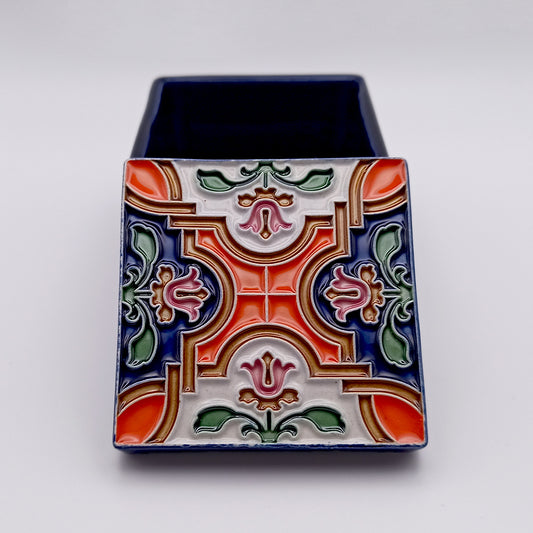 This Porto Covo Ceramic Box by Tejo Shop is colorful and hand-painted with intricate floral and geometric patterns in red, green, blue, and pink on the lid. The box itself is a deep blue, creating a striking contrast with the vibrant design on the top. Perfect as an elegant ceramic décor piece, it features a plain white background.