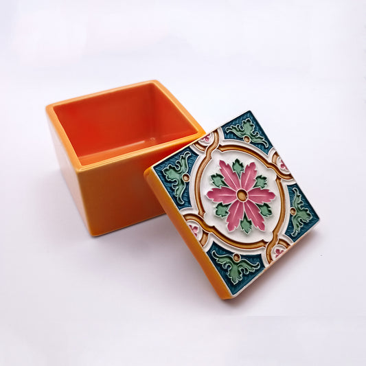 A small, square, Porto Santo Island Ceramic Box by Tejo Shop in luxurious orange ceramic with an ornate, colorful lid featuring a central pink flower and decorative green, blue, and white patterns is open, revealing the empty, handcrafted orange interior.