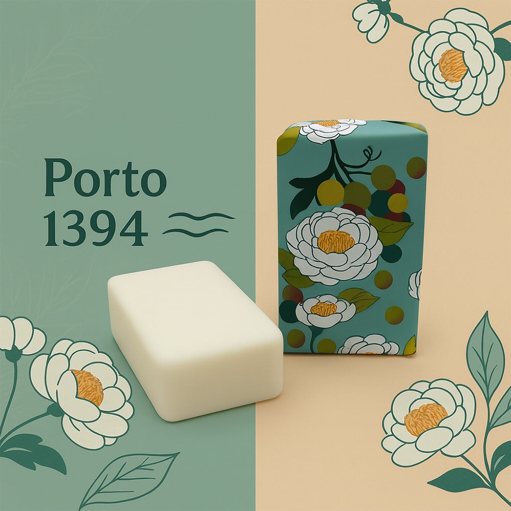 Rectangular Porto 1394 – Birthplace of the Navigators soap bar by 100ml with camellia scent sits beside its floral-patterned box on a green and beige background. White and yellow flowers, "Porto 1394" text, and wavy lines highlight soap-making heritage.