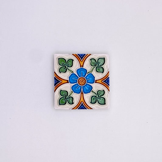 A decorative ceramic tile featuring a colorful floral design with blue, green, orange, and white patterns centered around a blue dot on a gray background, reminiscent of Tejo Shop's Spanish Style Tiles.
