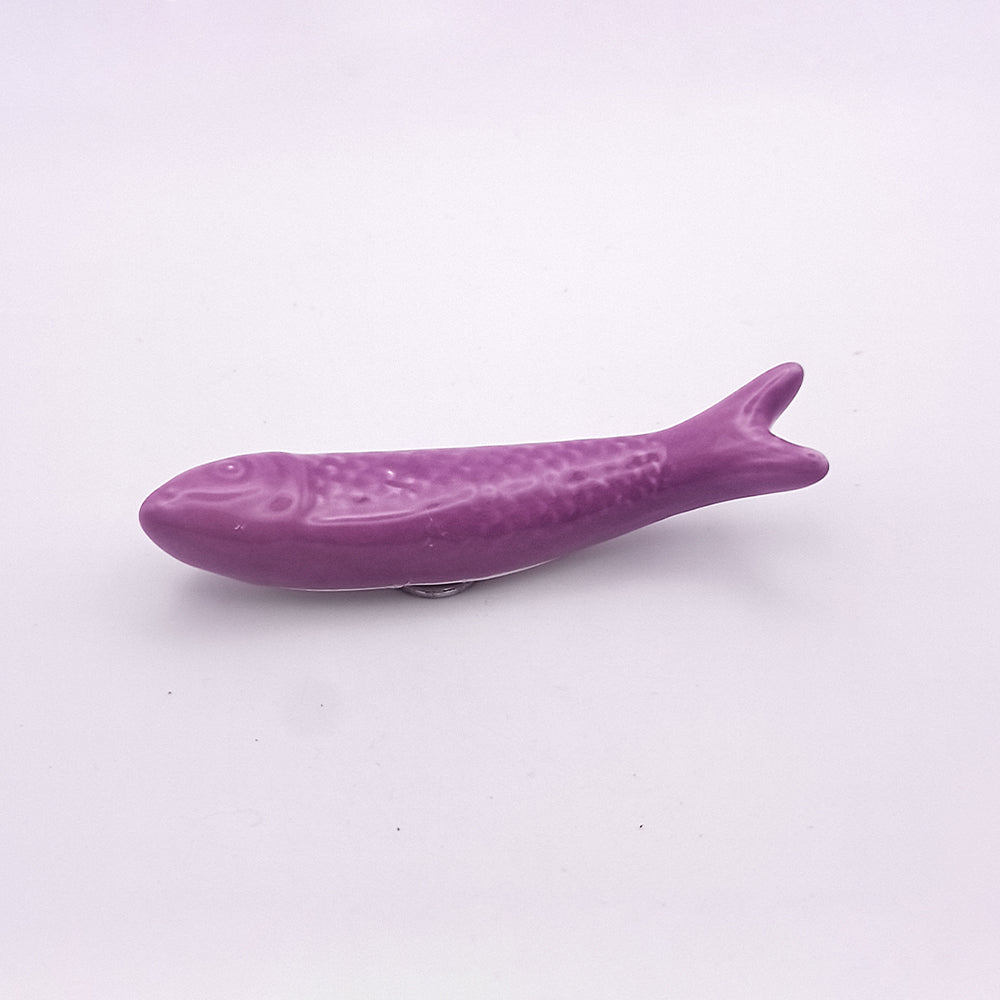 A small, purple, fish-shaped object is placed against a plain white background. The Tejo Shop Ceramic Sardine Fridge Magnet has a smooth texture with detailing to represent scales and fins, making it a charming piece of kitchen decor and a true Portuguese icon.