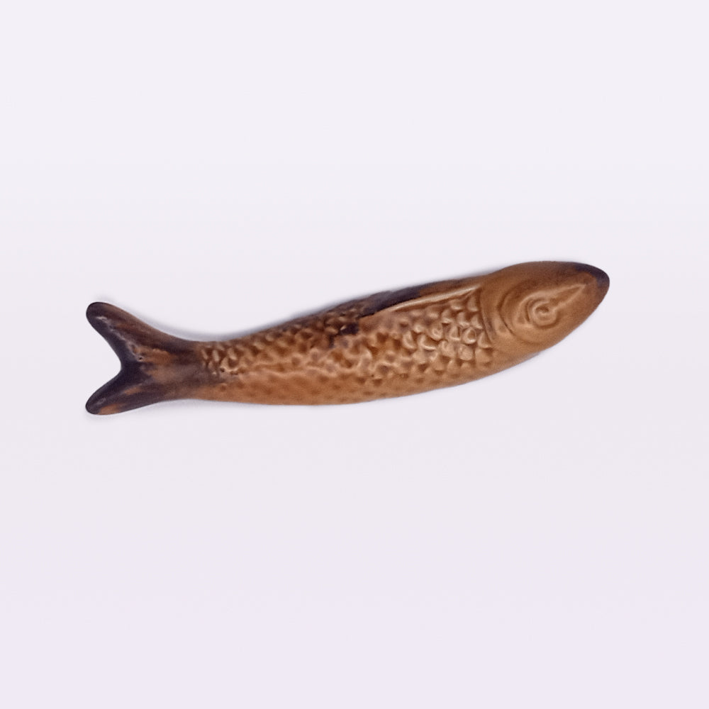 A small, brown ceramic fish with detailed scales and fins lies flat on a plain white background. This fish, reminiscent of the Ceramic Sardine Fridge Magnet by Tejo Shop, is elongated with a rounded head and pointed tail—perfect for adding charm to your kitchen decor.