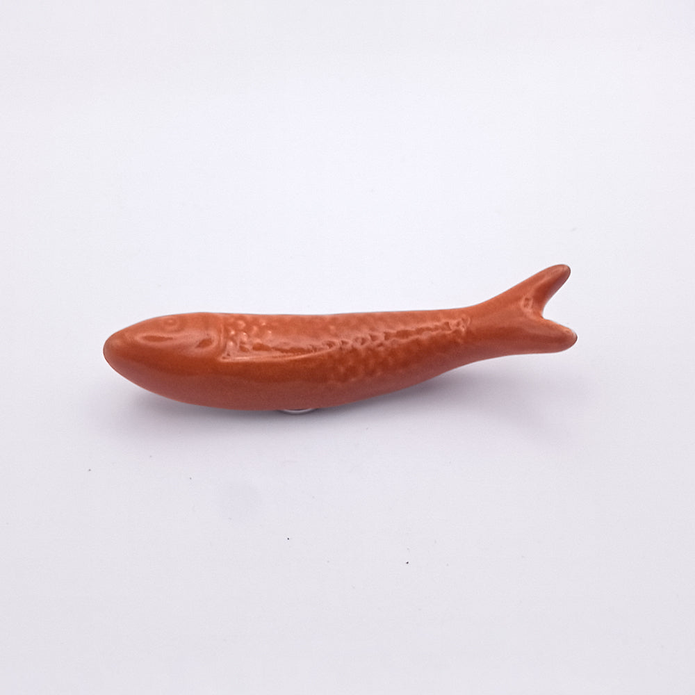 A small, smooth, brown ceramic object shaped like a fish with textured details on its body and fins rests flat on a plain white surface, perfectly capturing the essence of a Portuguese icon. This charming piece from Tejo Shop doubles as their Ceramic Sardine Fridge Magnet, making it ideal for stylish kitchen decor.