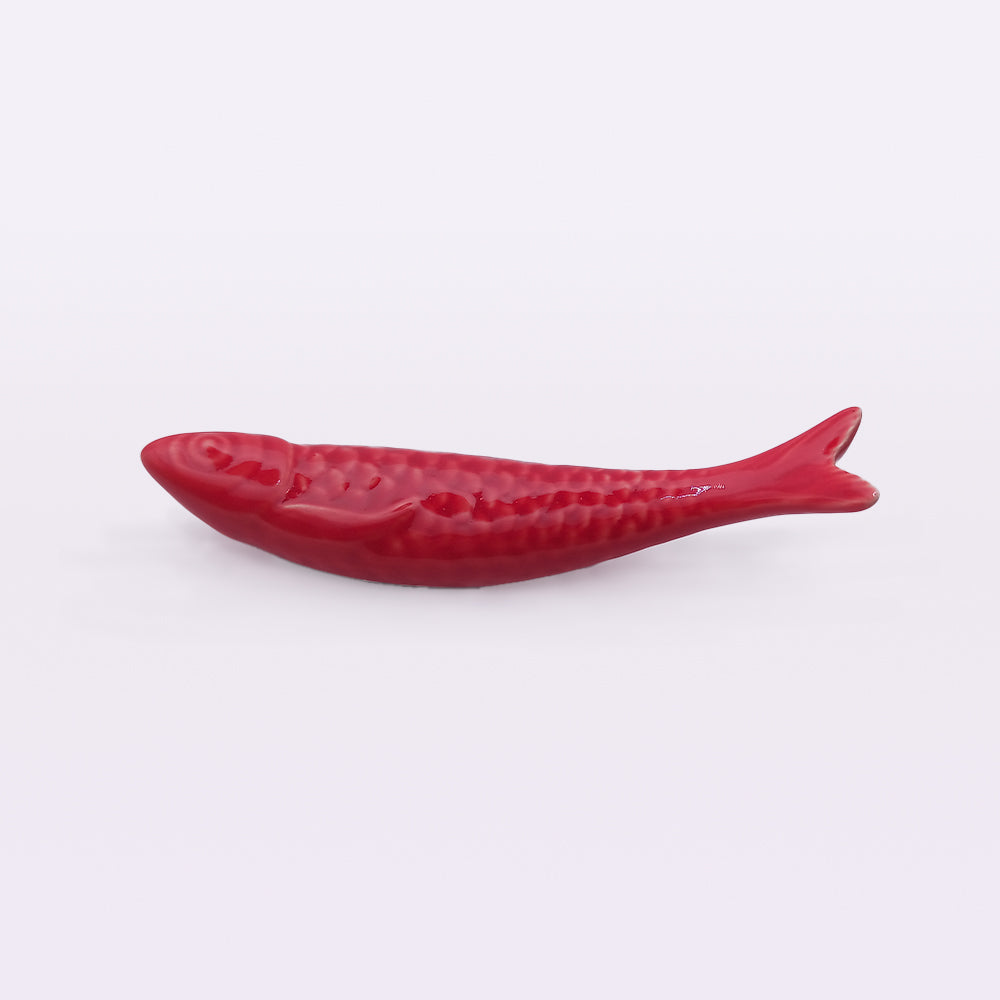 A red, fish-shaped object lying on a white surface. The handcrafted piece appears to have a smooth, glossy texture with detailed scales and fins, resembling Portuguese Ceramic Sardines from Tejo Shop and making for a unique addition to home decor.