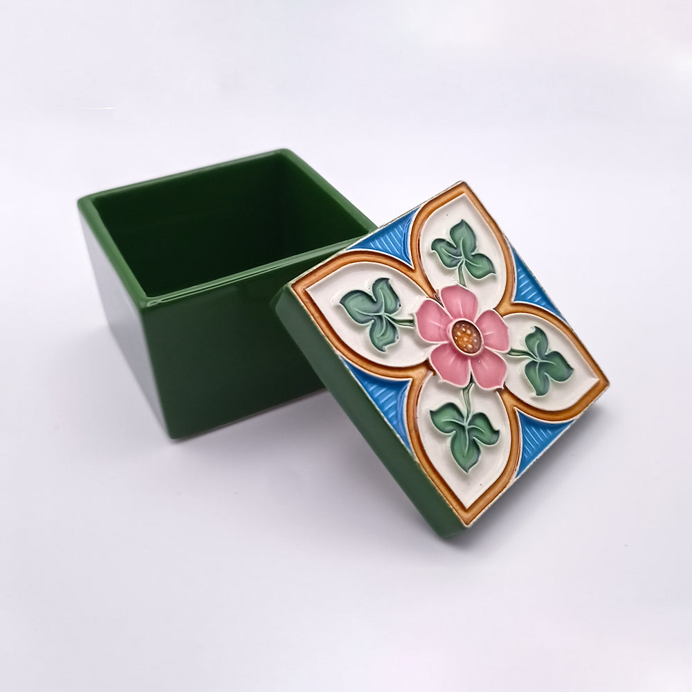 A small, square, green Sagres Ceramic Box by Tejo Shop with a removable lid. The luxurious lid features a colorful floral design with a pink flower at the center, surrounded by green leaves and blue geometric patterns set against a white background. The hand-decorated box is open, revealing an empty interior.