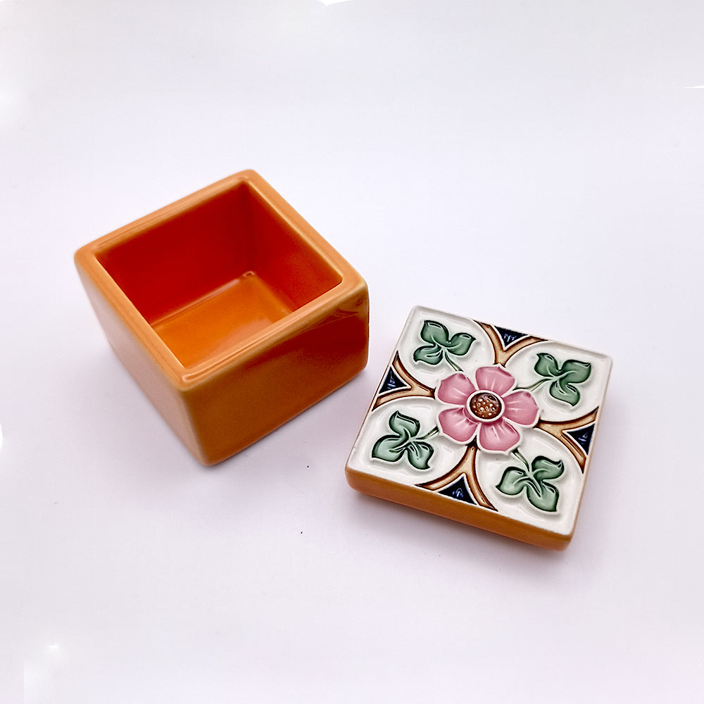 The Sintra Ceramic Box by Tejo Shop is a small square ceramic piece with an orange base and a detachable lid. The lid features a colorful floral design with a pink flower at the center, surrounded by green leaves and a decorative pattern, exuding handcrafted artistic charm. The box is open, showing its empty interior.