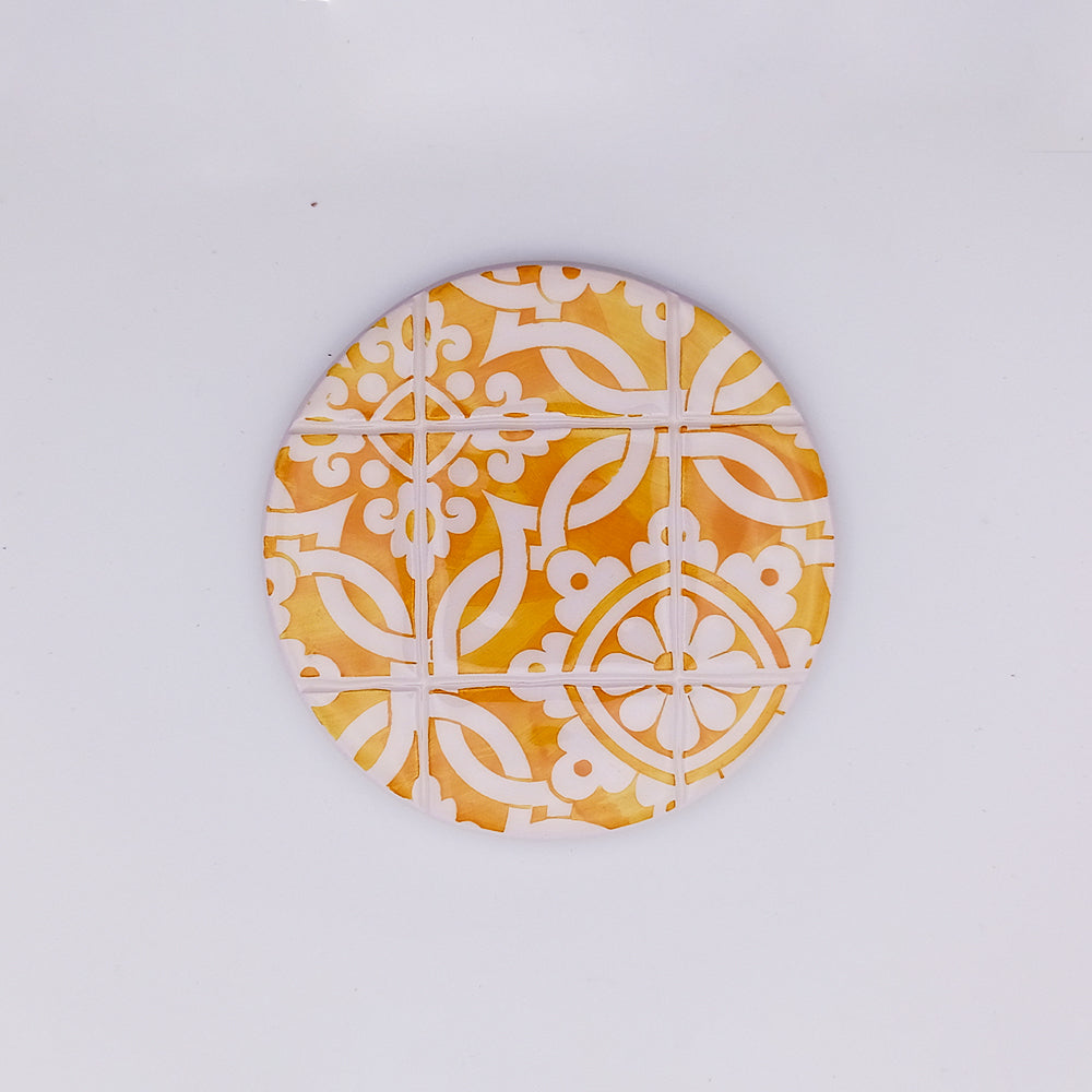 A round, hand-made coaster featuring a geometric pattern in white and varying shades of yellow and orange. The design, reminiscent of a traditional Sintra Ceramic Cup Pads by Tejo Shop, consists of symmetrically arranged curved and angular shapes on a plain white background.