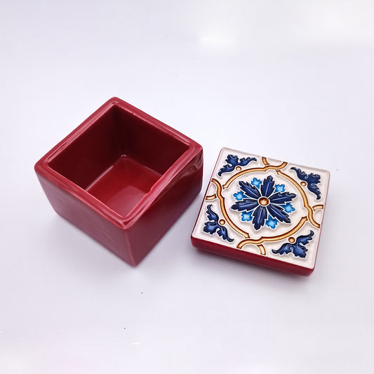 A small, red square ceramic box with its lid off showcases superb craftsmanship. The lid is decorated with a blue, white, and gold floral pattern. The interior of the Terceira Island Ceramic Box by Tejo Shop is plain red and empty. The photo is taken on a white background.