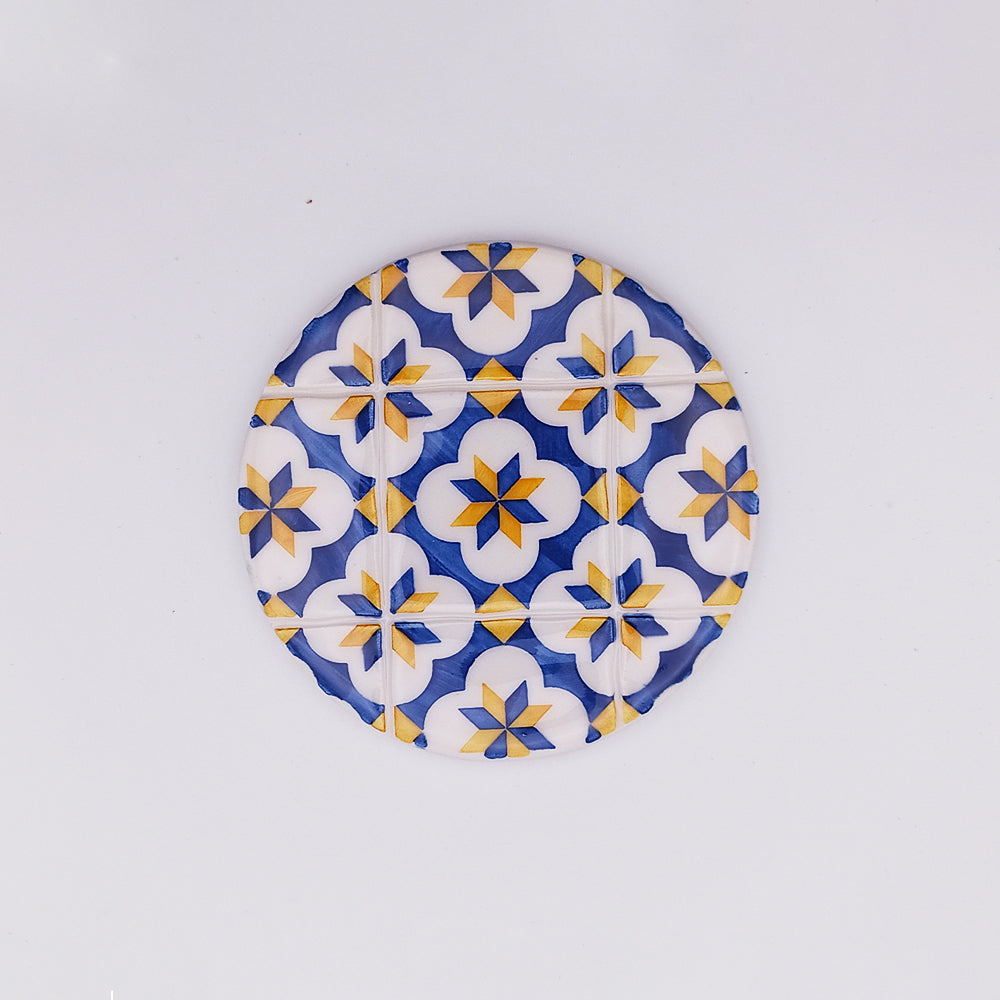 A round Vilamora Ceramic Tile Cup Pad by Tejo Shop with a vibrant geometric pattern featuring white, blue, and yellow stars and diamonds. The design includes floral motifs and symmetrical shapes, creating an intricate, tile-like appearance. The background is plain white.
