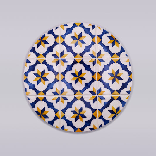 A circular Vilamoura Ceramic Hot Plate Trivet by Tejo Shop design featuring a repeated pattern of blue, yellow, and white geometric shapes. The hand-made trivet includes star-like motifs enclosed within white floral shapes, set against a blue background, creating an intricate and symmetrical visual effect with artisanal flair.
