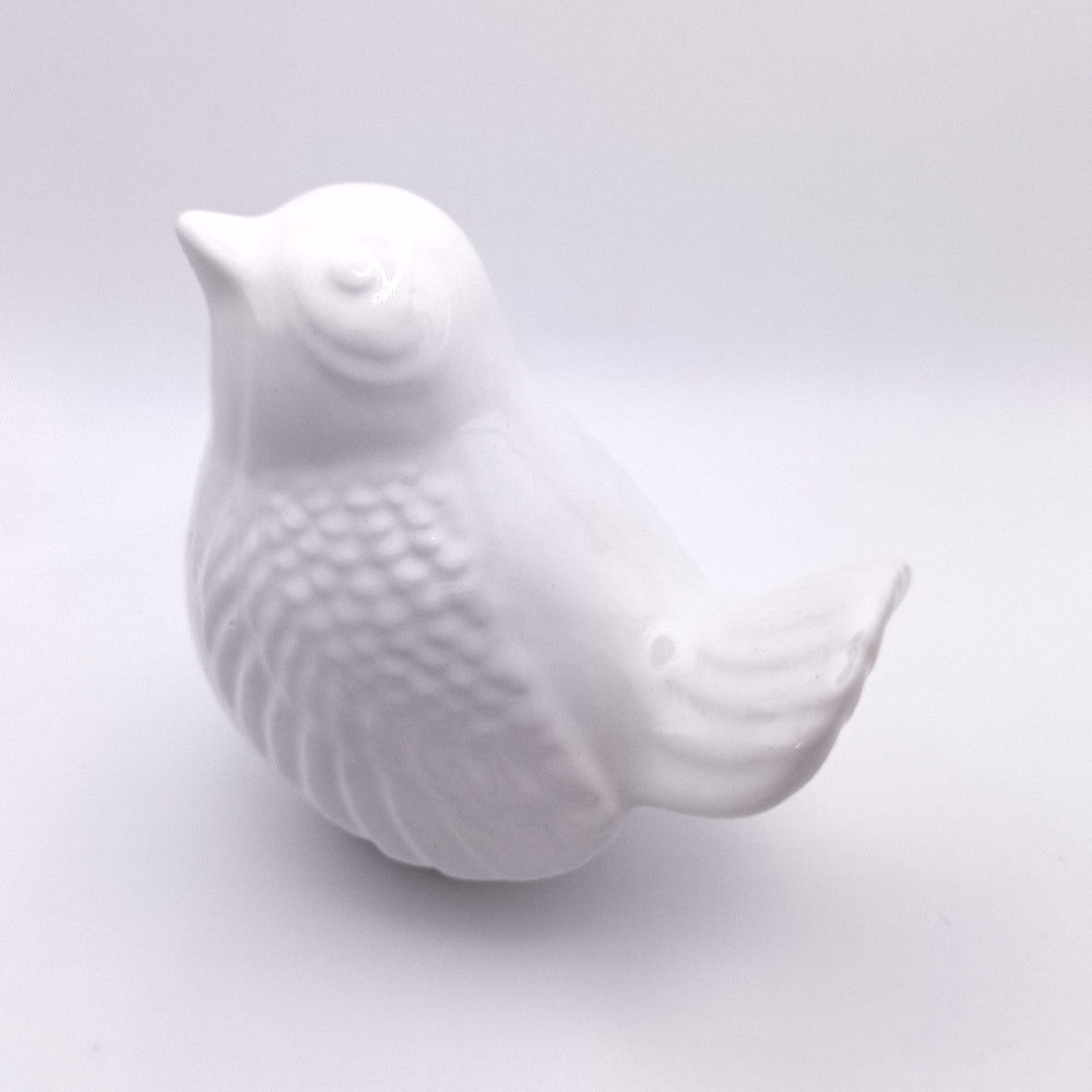 A handmade white ceramic bird figurine from Tejo Shop, known as the "Ceramic Bird looking up Figurine," is shown. This classy addition to any home decor is facing left with its beak slightly raised and eyes gently closed. The overall design is minimalistic and elegant, featuring a smooth, glossy surface with textured feathers.