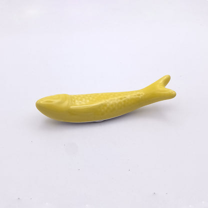 A small, yellow, fish-shaped object is centered on a plain white background. The Tejo Shop Ceramic Sardine Fridge Magnet features a smooth texture and minimal detailing, with subtle scales and a simple fish form, making it an ideal piece of kitchen decor reminiscent of a Portuguese icon.